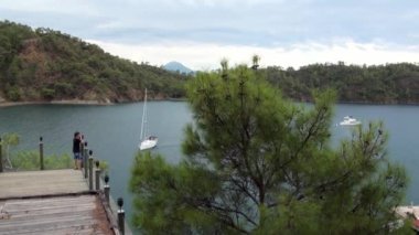 Çocuk köprüde duruyor ve sahildeki yat ve dağlara bakıyor..