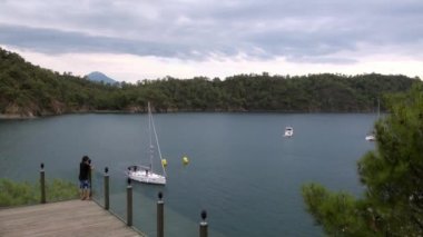 Çocuk köprüde duruyor ve sahildeki yat ve dağlara bakıyor..