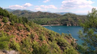 Beyaz yatlar arka planda yelken açıyor ve kıyı şeridinde dağlar..