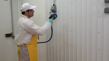 Erkek işçi endüstriyel atölyede beyaz duvarı yıkıyor ve dezenfekte ediyor.