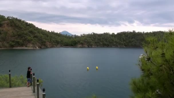 Garçon se tient sur le pont et regarde les yachts et les montagnes sur la côte dans la baie. — Video