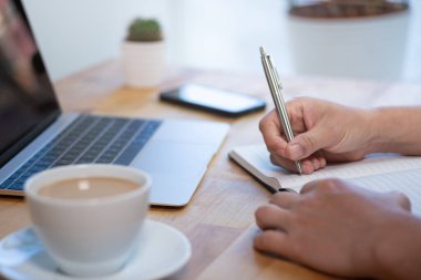 Woman writing in notepad. Working in office. Close up, shallow depth of field. Wooden desk with laptop, smartphone and coffee.