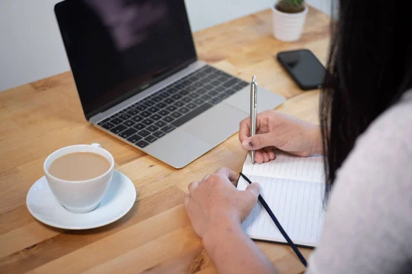 金属ペンでメモ帳に書いて働く女性 ノートパソコン コーヒー スマートフォン 植物とオフィスデスク — ストック写真