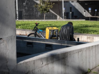 Çocuk eve yemek getiriyor, dışarıda dinleniyor, küp şeklinde sırt çantasının yanında oturuyor..