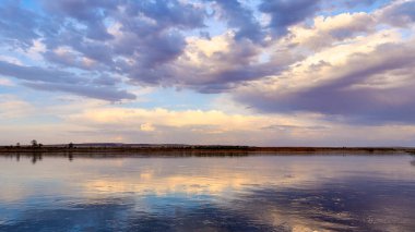 Sakin bir akıntıyla geniş bozkır nehrinin panoramik manzarası ve suya yansıyan dramatik bulutlarla dolu gökyüzü.