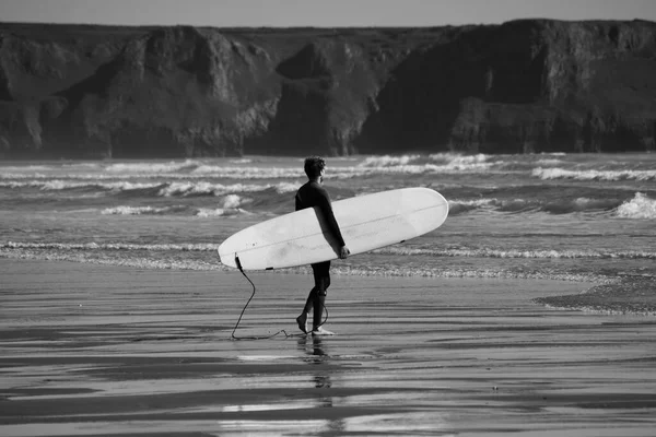 Surfers of all ages carry their surfboards to the water's edge of Llangennith beach on the Gower Peninsula. Monochrome image depicting outdoor activities, positive and healthy lifestyles.