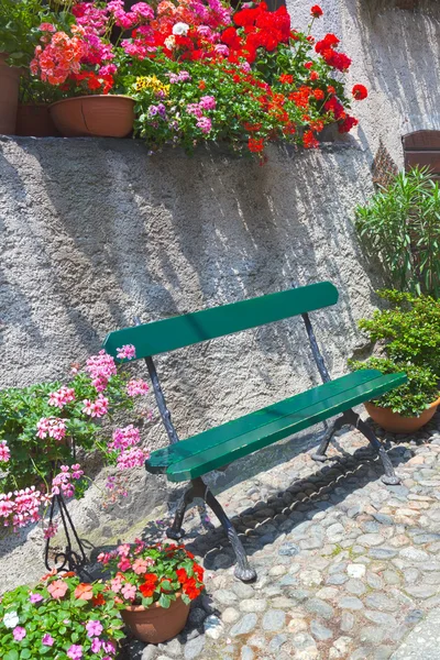 Green bench on cobbled street by old stone wall, surrounded by hanging geranium plants and flowers in pots — Stock Photo, Image