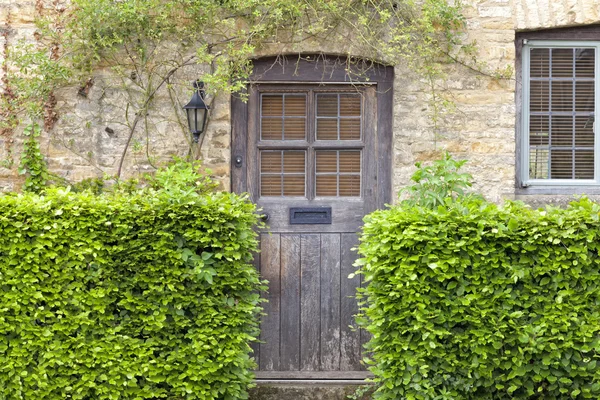 Eski kahverengi ahşap kapı geleneksel bal tarak ile yeşil kayın hedge önde, taş yazlık kırsal Cotswold köyde