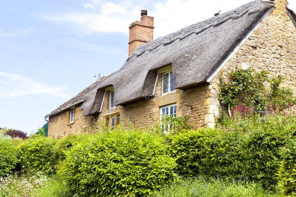 Traditional old Cotswold stone cottage with thatched roof and front garden overgrown with green shrubs on a sunny summer day