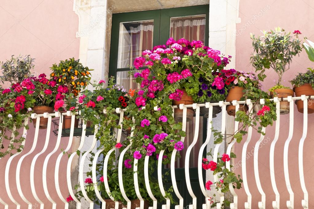 White metal balcony with floral display of colourful hanging plants, small olive, orange trees in an old stone building painted in pastel pink