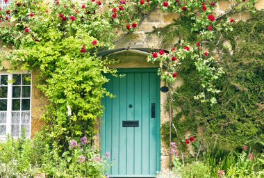 Green wooden doors in an old traditional English stone cottage surrounded by climbing red roses