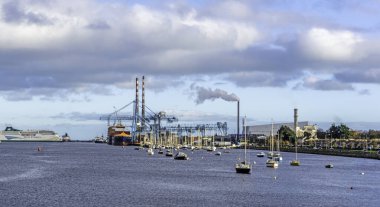 A view of Dublin Port, Ireland, with a gantry crane in operation, a passenger ferry berthed,  small boats and the East Link Ferry Bridge and the new incinerator in the distance.  clipart