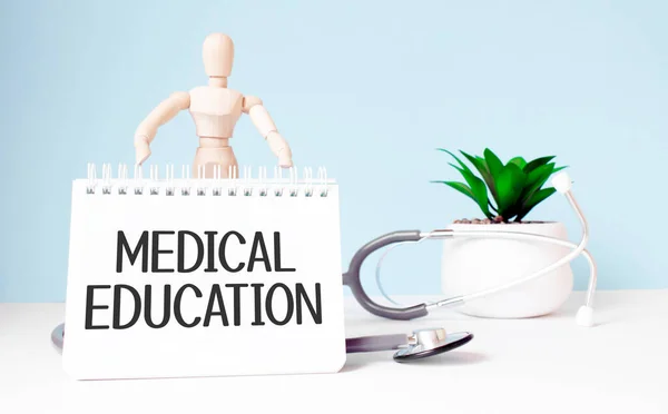 The text MEDICAL EDUCATION is written on notepad and wood man toy near a stethoscope on a blue background. Medical concept