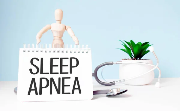 The text SLEEP APNEA is written on notepad and wood man toy near a stethoscope on a blue background. Medical concept