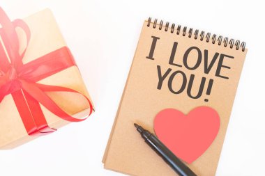 Sevgililer Günü konsepti. Kırmızı kurdeleli Creaft hediye kutusu, pembe ahşap kalp, siyah kalem ve el işi renkli not defteri.