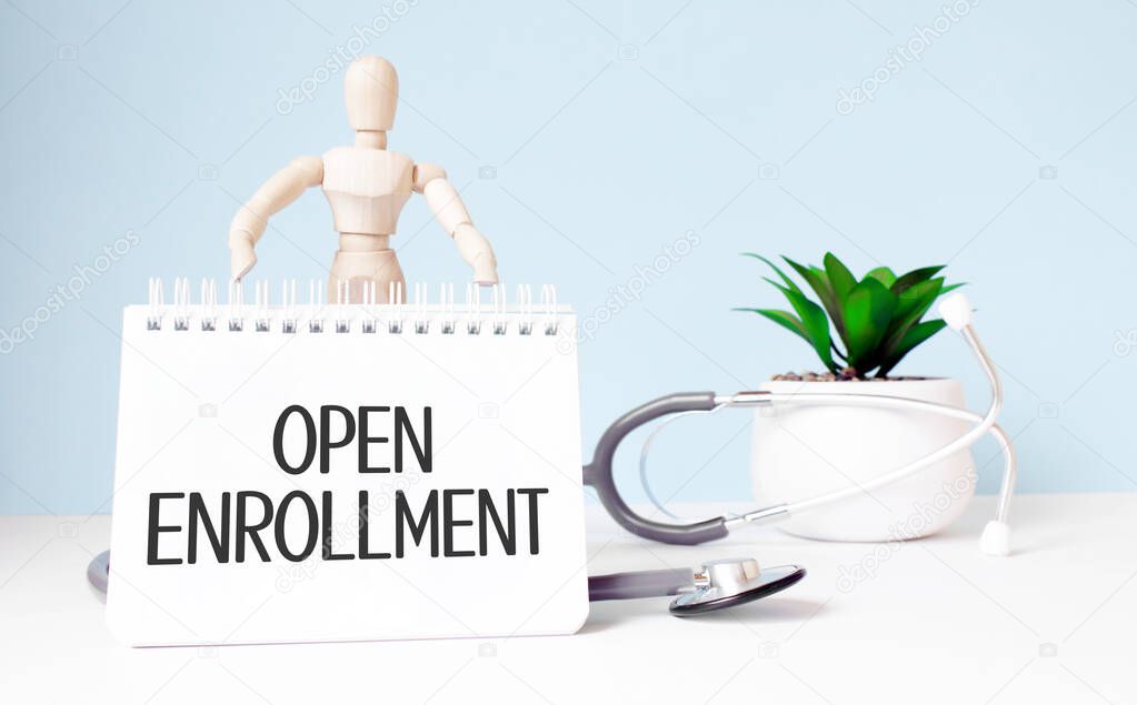 The text open enrollment is written on notepad and wood man toy near a stethoscope on a blue background. Medical concept