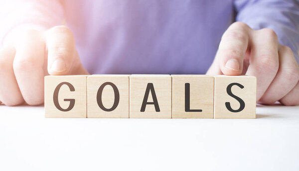 Бизнесмен держит в руках деревянный кубик с деловым словом GOALS на фоне стола. Миссия, видение и концепция основных ценностей