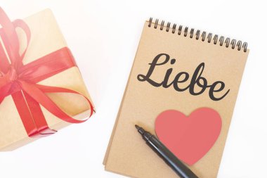 Sevgililer Günü konsepti. Kırmızı kurdeleli Creaft hediye kutusu, pembe tahta kalp, siyah kalem ve LIEBE işaretli el işi not defteri.