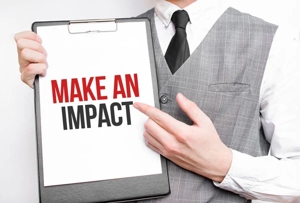 MAKE AN IMPACT inscription on a notebook in the hands of a businessman on a gray background, a man points with a finger to the text