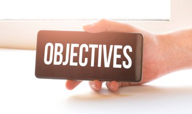 Adam evde kilit altında ve elinde OBJECTIVES imzalı siyah bir telefon tutuyor.