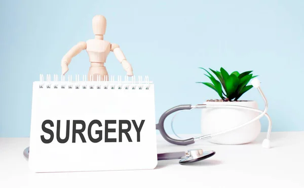 The text surgery, is written on notepad and wood man toy near a stethoscope on a blue background. Medical concept