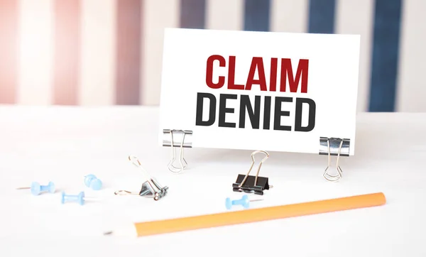CLAIM DENIED sign on paper on white desk with office tools. Blue and white background