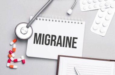 Tıp masasında MIGRAINE metni olan steteskop, hap ve defter..
