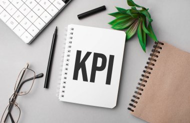 KPI, hesap makinesi, el işi renkli not defteri, bitki, siyah kalem ve gözlük içeren beyaz bir defterde yazılmıştır..