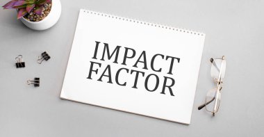 IMPACT FACTOR kalem, siyah çerçeveli gözlük ve yeşil bitkinin yanında beyaz bir deftere yazılmıştır..