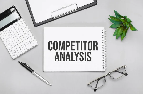 Writing text showing COMPETITOR ANALYSIS. Calculator,pen,plan,glasses and black folder on grey background