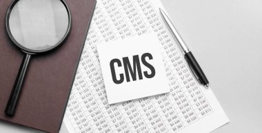 Büyüteç, finansal belge, CMS işareti ve gri arkaplanda kahverengi defter..