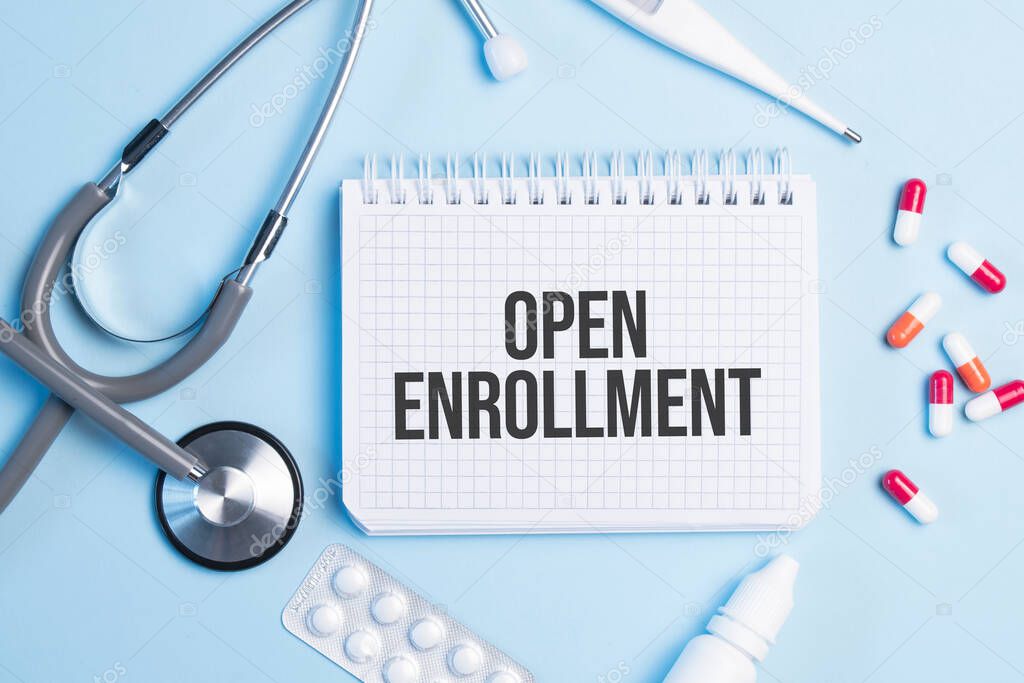 The word open enrollment written on a white notepad on a blue background near a stethoscope, syringe, electronic thermometer and pills. Medical concept