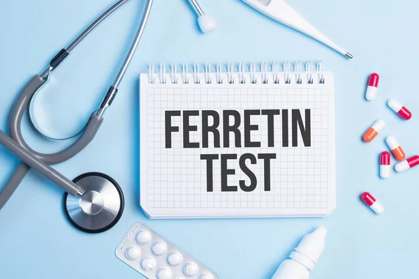 The word ferretin test written on a white notepad on a blue background near a stethoscope, syringe, electronic thermometer and pills. Medical concept