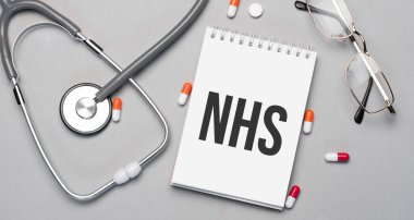 In the notebook is the text NHS, next to a stethoscope, pills and glasses.