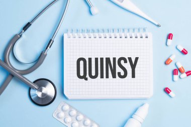 The word quinsy written on a white notepad on a blue background near a stethoscope, syringe, electronic thermometer and pills. Medical concept