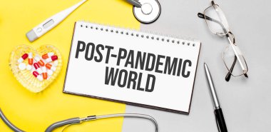 Text post-pandemic world on notebook with stethoscope,glasses, pen,thermometer, red pills and pen on yellow background. Medical concept.