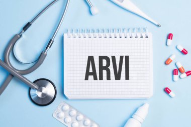 Stetoskop, şırınga, elektronik termometre ve hapların yanındaki mavi arka planda beyaz bir not defterine arvi yazıldı. Tıbbi konsept