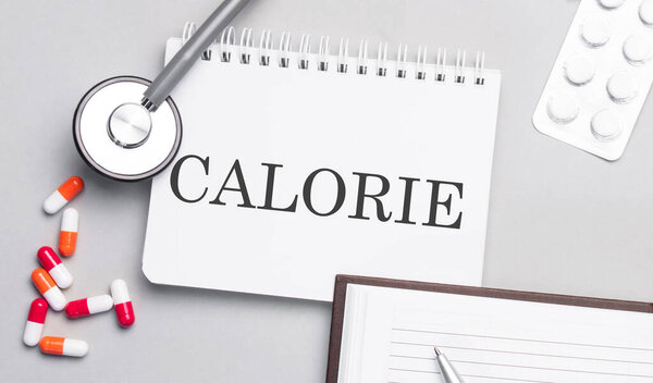 Стетоскоп, таблетки и блокнот с калорийным текстом на медицинском столе. CALORIE