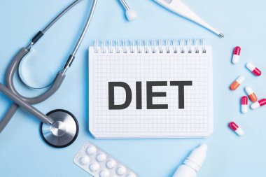 Diyet kelimesi beyaz bir not defterinde steteskop, şırınga, elektronik termometre ve hapların yanında mavi bir arkaplanda yazıyor. Tıbbi konsept