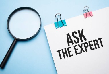 Magnifying glass loupe with sign Ask the Expert on the paper notepad wtih colored paper clips on the blue table.