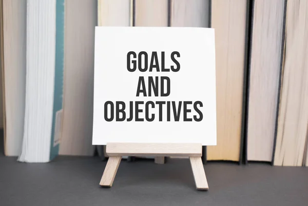 White card with text Goals and Objectives stands on the desk against the background of books stacked