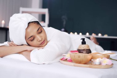 Beyaz bornoz ve başörtüsü takmış gülümseyen Asyalı kadın yatağa uzanıp Tayland 'daki alternatif tıp tedavi merkezindeki masaj terapisine hazırlanıyor.