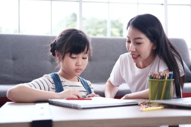 Ciddi Asyalı anne küçük kıza oturma odasında hayal gücü için tahta renkli kalemlerle resim yapmayı ve resim yapmayı öğretiyor. Homeschool ve eğitim konseptine sahip aile
