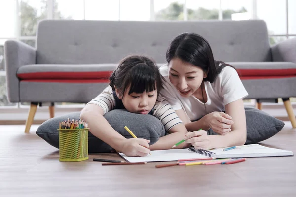 Smiling Asian mother and little asian girl child is drawing and Painting with wooden colored pencils on paper for imagination together in living room. Homeschool and educational concept