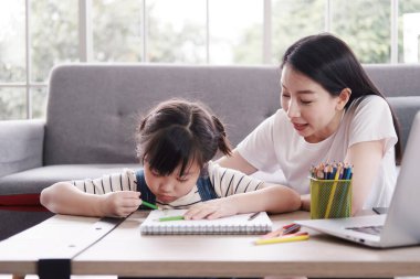 Gülümseyen Asyalı anne ve küçük kız çocuğu oturma odasında kağıt üzerine tahta renkli kalemlerle resim yapıyor. Bilgisayarla öğreniyor. Ev okulu ve eğitim konsepti