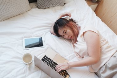 Gülümseyen Asyalı iş kadını dinleniyor ve tatilde yatak odasında dizüstü bilgisayarla yatıyor. Covod-19 salgını sırasında evde teknolojiyle çalışıyor..