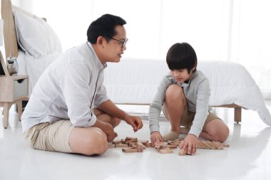 Gülümseyen Asyalı baba ve küçük tatlı çocuk tatilde yatak odasında tahta blok oyunu oynarken eğleniyor ve eğleniyorlar. Aile aktiviteleri ve evde eğitim kavramı için çocuk eğitimi.