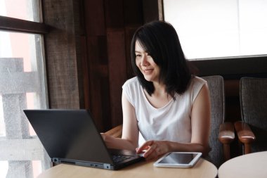 Gülümseyen Asyalı genç kadın internette çalışıyor ya da bilgisayarla alışveriş yapıyor. İnternetten bilgi topluyor ve evden çalışıyor. İş kadını teknoloji konseptiyle çalışıyor.