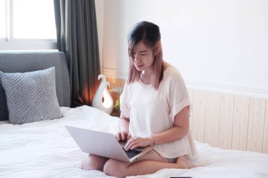 Gülümseyen Asyalı genç kadın internette çalışıyor ya da yatak odasında dizüstü bilgisayarla alışveriş yapıyor. İnternetten bilgi topluyor ve evden çalışıyor. İş kadını teknoloji konseptiyle çalışıyor.