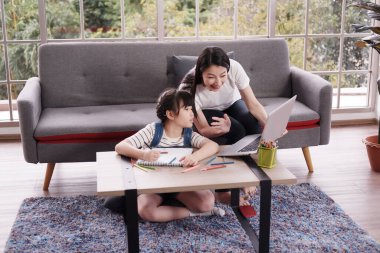 Gülümseyen Asyalı anne küçük kız çocuğuna ödev veriyor tahta renkli kalemlerle resim çiziyor. Bilgisayarla çocuk öğrenimi. Ev okulu ve eğitim konsepti